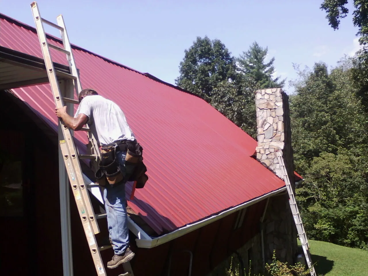 Roofer on a ladder beside a red metal roof and stone chimney — Soffit & Fascia Repair by Roofing Brothers in Madisonville